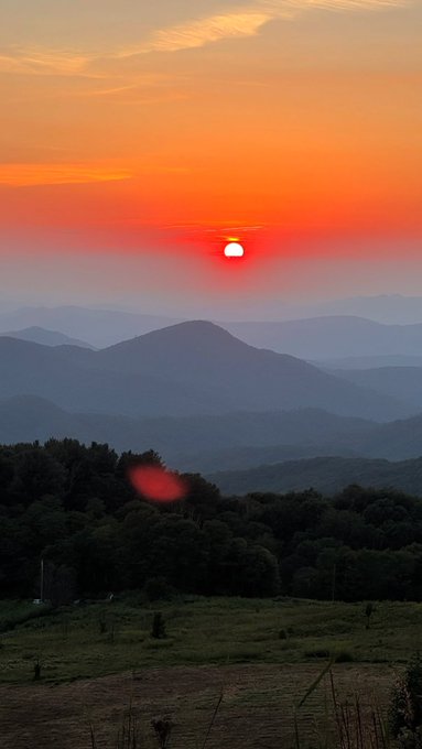 A scenic photograph of a vibrant orange sunset over a layered mountain range, with a dark forest in the foreground.