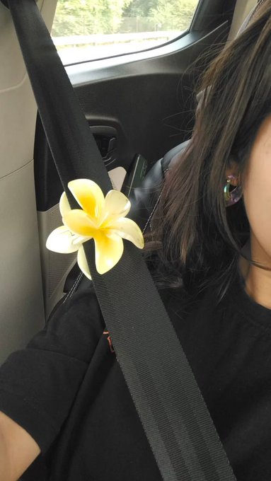 A selfie taken inside a car showing a woman with dark hair and an earring, with a yellow and white plumeria flower tucked into her black seatbelt.