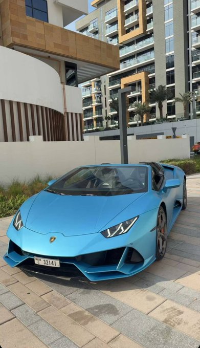 A sleek blue Lamborghini Huracán with a Dubai license plate (T 32141) is parked outside a modern high-rise residential building. The image perfectly embodies the post's "GM to everyone who says GM" flex—a casual greeting paired with a luxury supercar as the ultimate status statement.