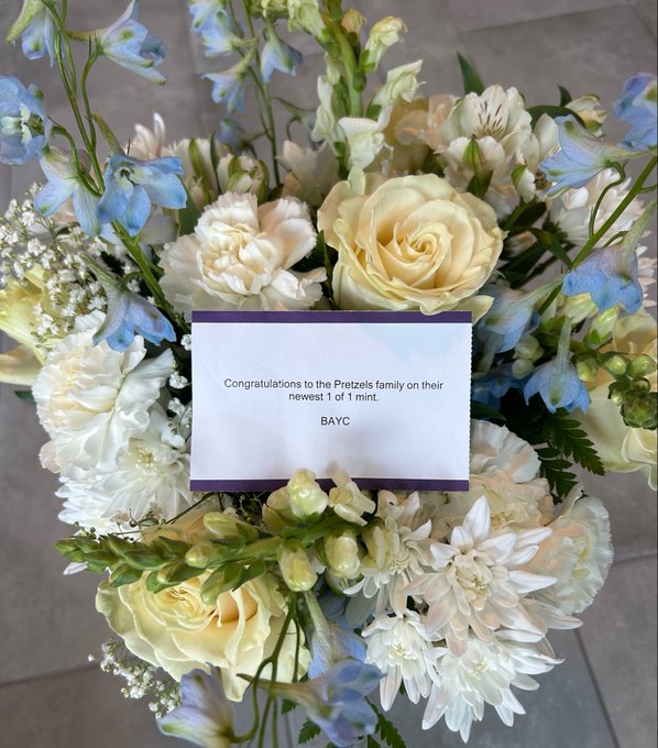 A floral arrangement of white, cream, and light blue flowers surrounds a card reading 'Congratulations to the Pretzels family on their newest 1 of 1 mint. BAYC'.
