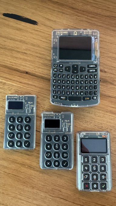 Four COLDCARD hardware cryptocurrency wallets of varying sizes, featuring transparent casings that reveal their internal circuit boards and full QWERTY or numeric keypads, are arranged on a wooden surface in preparation for a cold storage workshop.