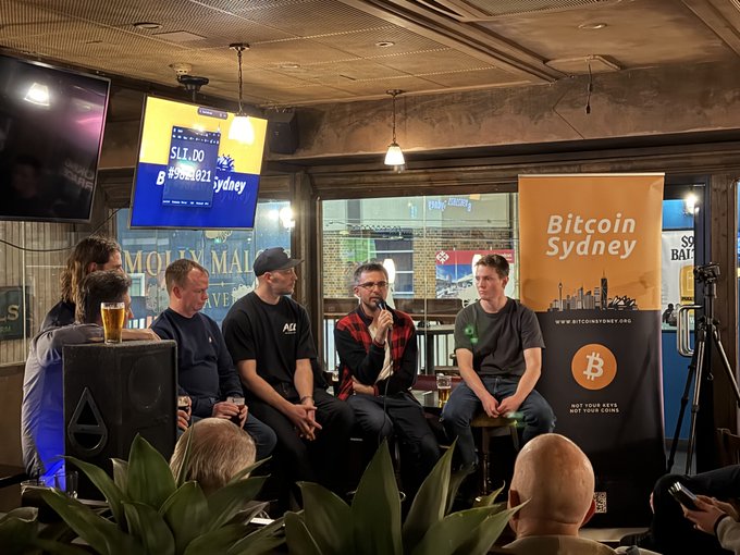 A panel of speakers sits on stools at a Bitcoin Sydney meetup, with one man speaking into a microphone while others listen, framed by a Bitcoin Sydney banner and a screen displaying "$11.00" and "B #308827". The setting matches the post perfectly, capturing the community gathering hosted by the Bitcoin Sydney group.