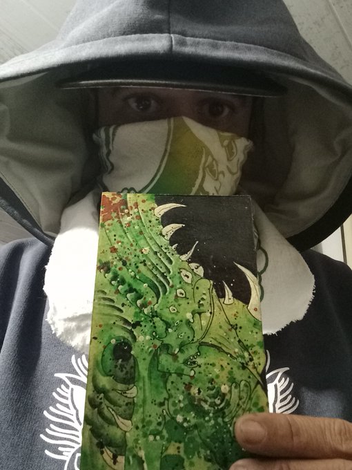 A person wearing a hooded jacket and a patterned face mask holds up a green-toned fantasy illustration card, with only their eyes visible above the mask.