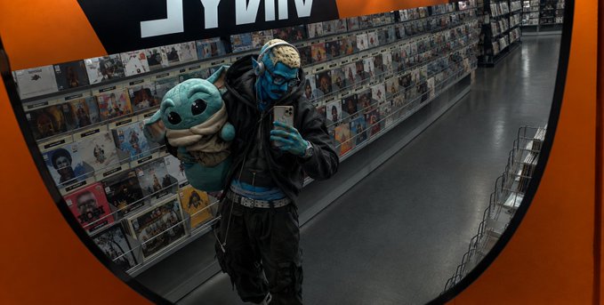A person in a denim outfit and blue alien-like makeup poses with a plush toy while taking a mirror selfie inside a vinyl record store, with rows of CDs visible behind them.
