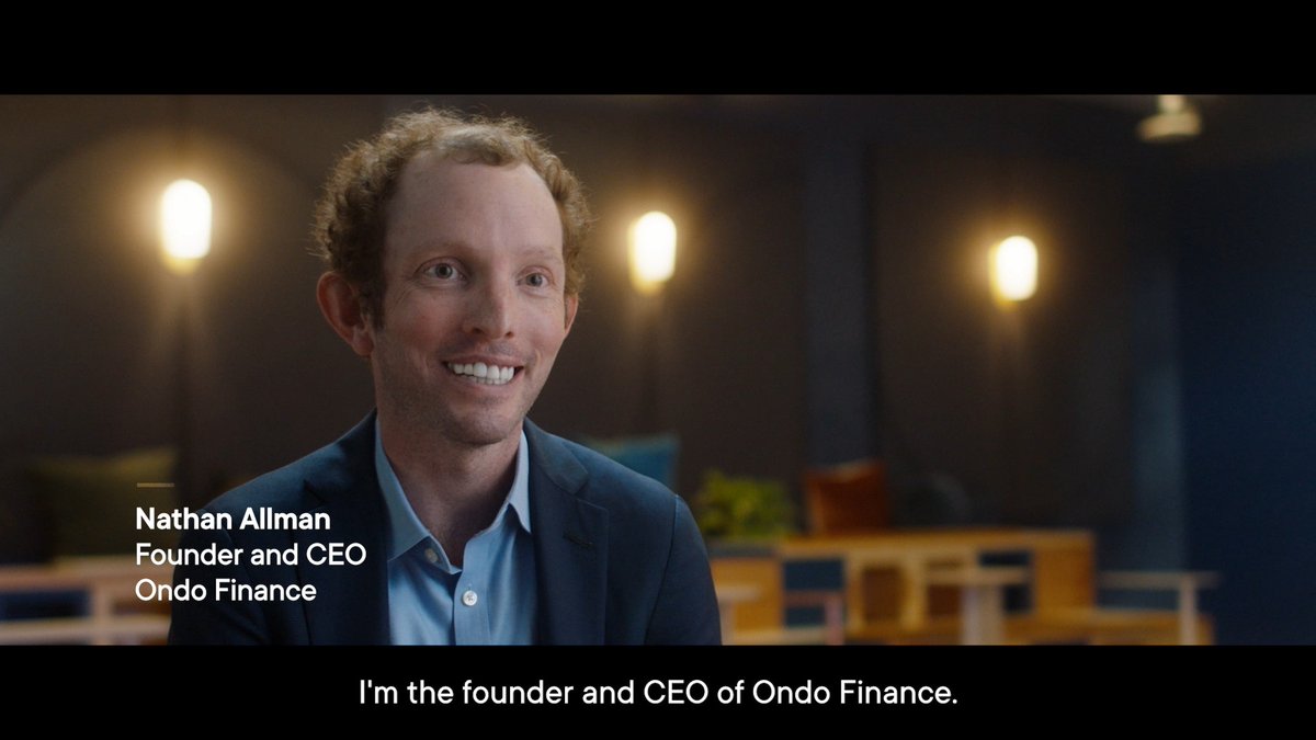 The image features a man wearing a suit, smiling at the camera while sitting in front of a blue wall with three lights behind him. The text "Sad to hear about the passing of Nathan Allman" is written below his photo. This suggests that the image may be related to an event or news story involving Nathan Allman and the Ondo + Ripple collaboration, which focuses on real-world asset tokenization.