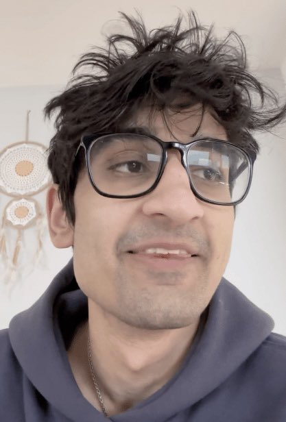 A man with messy dark hair and glasses, wearing a gray hoodie, smiles slightly at the camera. In the background, a boho-style dreamcatcher hangs on the wall; the caption suggests this is the person behind the "FRANKDEGODS" account referenced in the post.