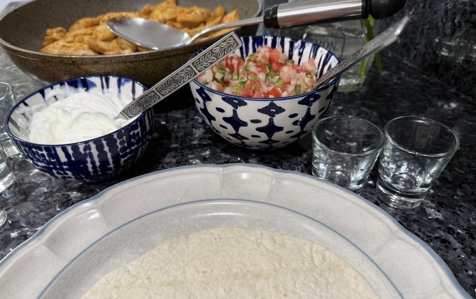 Photograph of food preparation on a dark marble countertop, showing bowls of sour cream, fresh tomato and onion salsa, fried items, two small shot glasses, and a large container of dough in the foreground.