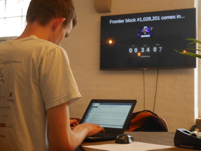 A person wearing a light t-shirt works on a laptop while a wall-mounted TV displays a countdown timer for "Frontier block #1,028,201" at 00:34:07.