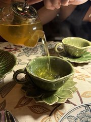 A hand pours steaming green tea from a glass pot into a leaf-shaped ceramic cup, with a matching leaf-shaped saucer on a decorative placemat.