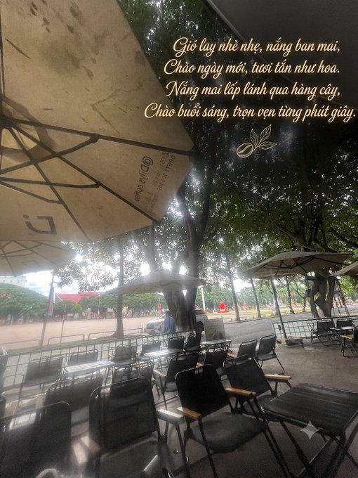 View from under a café umbrella at an outdoor seating area with empty chairs and tables, overlaid with Vietnamese text that reads: 'Gió lay nhẹ nhè, nâng bàn mai, Chào ngày mới, tươi tân như hoa. Nắng mai lấp lánh qua hàng cây, Chào buổi sáng, trọn vẹn từng phút giây.'