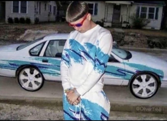The image features a man wearing a blue and white outfit standing next to a car with blue and green designs painted on its hood, which appears to be a luxury car. The surrounding environment consists of houses and trees along with a sidewalk. The main focus of the image is the man in the outfit posing near the car.