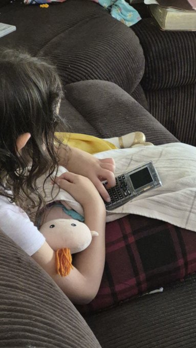 A young person with long hair lying on a brown couch, holding an old mobile phone while cuddling with a stuffed toy.