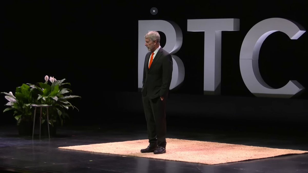 The image features a man standing on a stage with a rug beneath him. The man appears to be dressed in a suit, possibly a black suit, and he has his hands in his pockets. In front of him, there's a potted plant that adds some greenery to the scene. The background is dark, which contrasts with the bright colors of the man's suit and the potted plant. There are also words written on the wall behind the man, including "bitcoin", "LUNC A", and "@saylor starts buying $BTC". These words seem to be related to a post or message about Bitcoin, possibly discussing its decentralized nature and the potential impact of @saylor's actions in the cryptocurrency world.