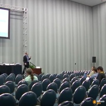 A speaker presents at a podium in a large conference hall with rows of blue chairs and a Bitcoin logo visible in the corner.
