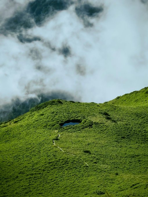 A lush green mountainside is partially shrouded in misty clouds, with a small pond visible on the slope.