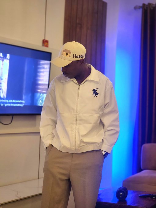 A man in a white “Hoedat” cap and white jacket stands with his hands in his pockets beside a TV and blue accent lighting.