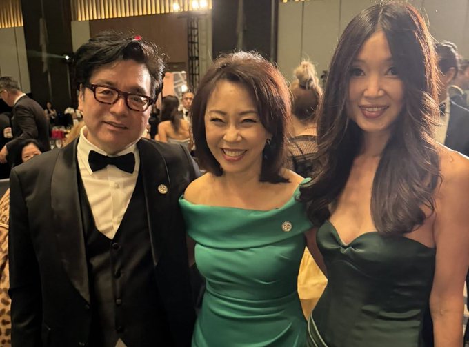 Three well-dressed individuals—a man in a tuxedo, a woman in an emerald green dress, and a younger woman in a dark green dress—smile together at what appears to be a formal gala event.