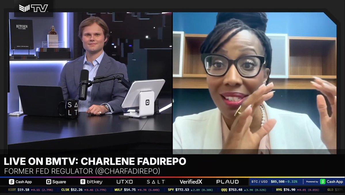 A screenshot from BMTV showing an interview titled 'LIVE ON BMTV: CHARLENE FADIREPO, FORMER FED REGULATOR (@CHARFADIREPO)' with a male host on the left and a woman with glasses speaking on the right, above a stock ticker displaying BTC/USD at $85,508.