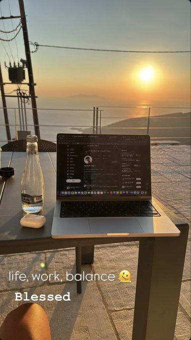 A laptop sits on an outdoor table overlooking a sunset over the sea, with a beverage bottle nearby, captioned 'life, work, balance Blessed' with a smiley emoji.