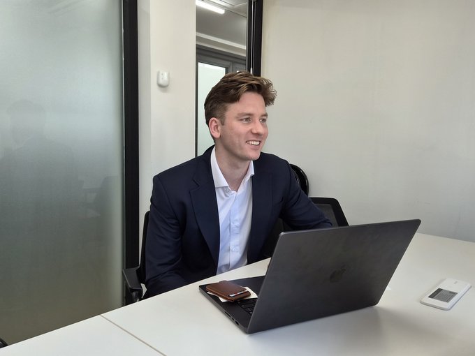 A smiling man in a navy blazer and white shirt sits at a desk with an open laptop during an office interview setting. The image illustrates the article's caption, showing the co-founder engaged in a discussion with Yonhap News about institutional Ethereum adoption in South Korea.