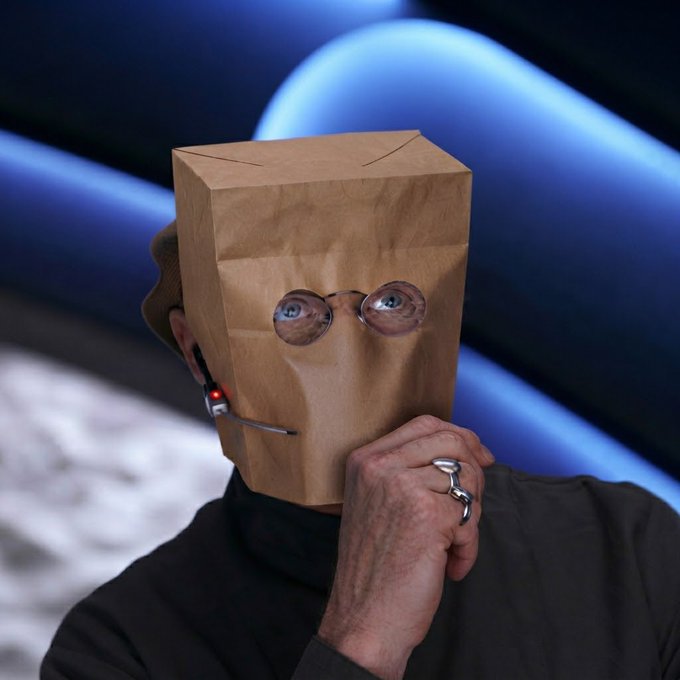 A person wearing a brown paper bag over their head with cutout eye holes and glasses peers out in a contemplative pose, resting their chin on a hand adorned with silver rings, while wearing a headset and dark clothing against a blue background—evoking the bewildered, hidden identity of someone trying to make sense of $zcat's wild, manipulative price swings from $170M down to $65M before a sudden pump to $300M.
