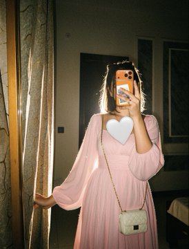 The image features a woman taking a selfie with her cell phone held up against a mirror. She is wearing a pink dress that stands out against the background. The room has curtains covering a window behind her, adding depth to the scene. A handbag can be seen in the foreground of the photo, and there are two beds visible in the background.