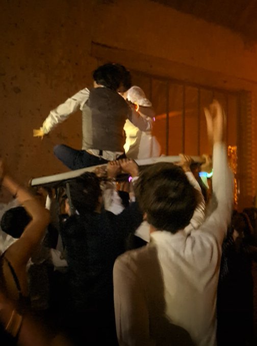 A man is being crowd-surfed on a chair, while another man in a white shirt raises his arms at what appears to be a lively wedding reception. The image captures the joyful, celebratory atmosphere referenced in the post about attending a French wedding and giving a speech in French.