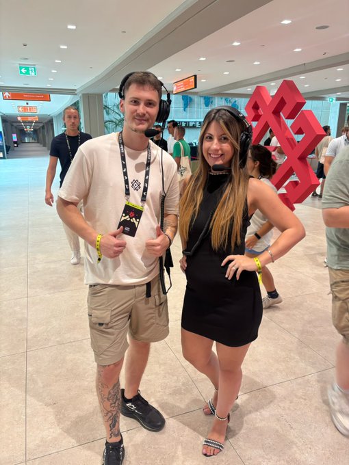 Two attendees wearing headsets pose together at the Solana Summit in Belgrade, with red "XX" Solana logo sculptures visible in the background of the convention hall. Both wear event lanyards and wristbands, capturing the social atmosphere of the crypto conference.