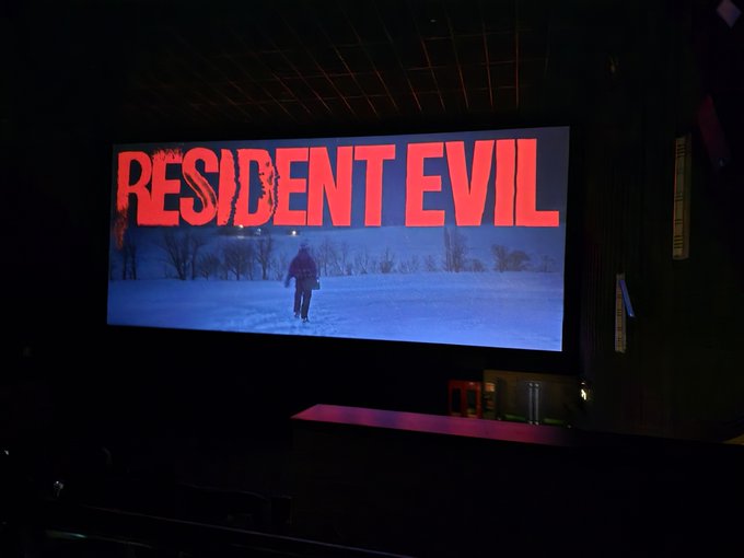 A movie theater screen displays the title "RESIDENT EVIL" in bold red letters above a snowy scene of a person walking through a forest.
