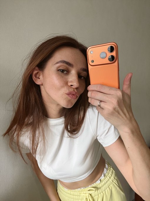 A young woman takes a mirror selfie wearing a white t-shirt and yellow pants while holding up an orange smartphone with a distinctive large camera module.