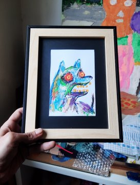 A hand holds a small framed colorful sketch of a fox, with art supplies and a child's painting visible in the background.