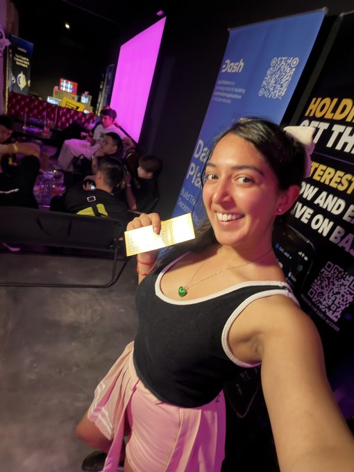 A smiling young woman in a black tank top and pink skirt takes a selfie while holding up a small ticket at a crowded event, with a 'Dash' promotional banner visible in the background.