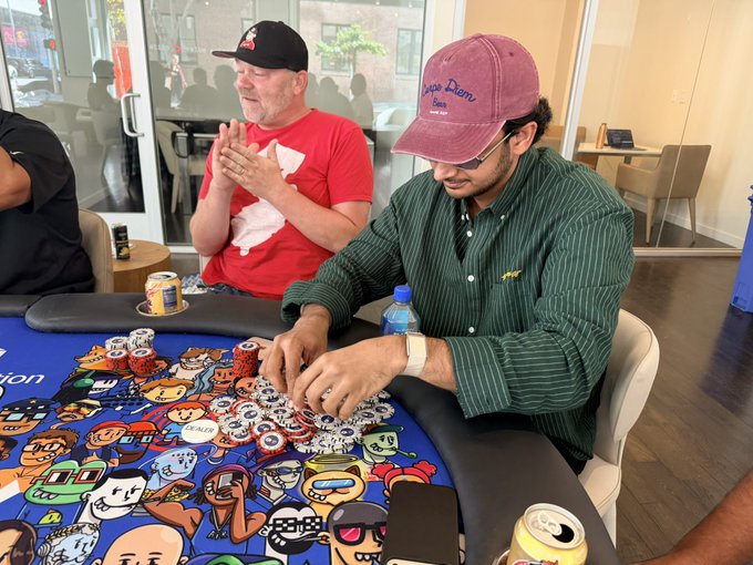 Two men sitting at a poker table; one in a red shirt is clapping, while another wearing a green striped shirt and a 'Pookie & Dianne' cap is focused on stacking chips at a colorful cartoon-themed poker table.