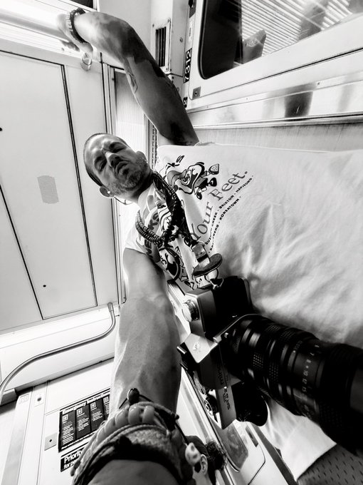 A black and white photo of a photographer on a train, holding a large camera with a telephoto lens, wearing a white graphic t-shirt that reads "Our Feet... serve... prioritize...".