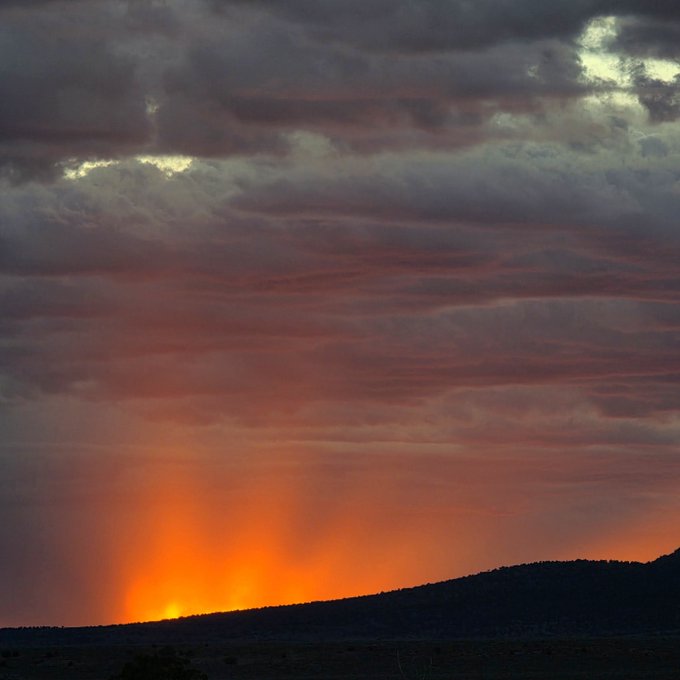 A dramatic landscape photograph of a sunset over silhouetted mountains, with dark clouds overhead and orange light streaming through the sky.