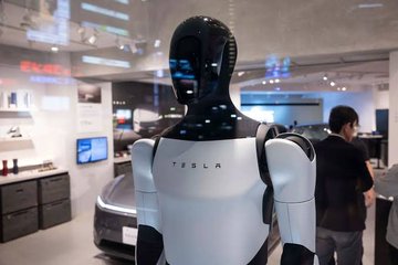 A Tesla humanoid robot is displayed at a showroom, with the "TESLA" logo prominently visible on its white chest plate, connecting to the post's mention of Elon Musk's work in physical AI. The sleek black-and-white robot stands in what appears to be a Tesla retail space, visually representing the "physical intelligence of tomorrow" referenced in the accompanying text.