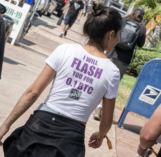 A woman seen from behind wearing a white t-shirt with purple text reading 'I WILL FLASH YOU FOR 0.1 BTC' above a QR code, walking on a boardwalk near a USPS mailbox.