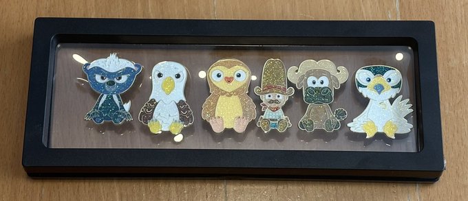 A display case holds six glittery VeeFriends pins featuring characters including a blue bird, a white bird, a yellow owl, a figure with a tall hat, a green monster, and a gray bird, showcased on a wooden surface. The collection aligns with the post, which advertises the seller's VeeFriends merchandise available for offers.
