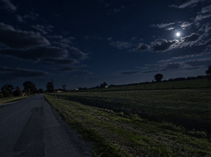 A nighttime landscape showing a country road beside an open field under a dark sky illuminated by the moon breaking through scattered clouds.
