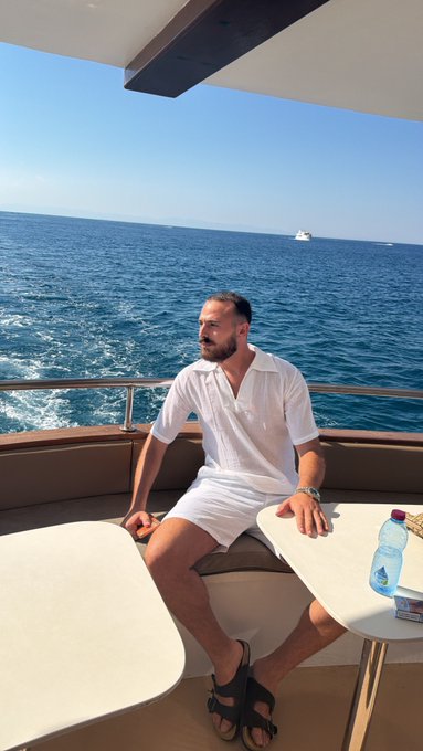 A relaxed man in a white linen outfit enjoys a morning on a boat, gazing out at the calm blue sea. The luxurious coastal setting perfectly captures the leisurely vibe of a "Gm friends" greeting.