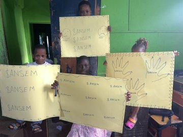 Children holding up handmade yellow signs repeatedly writing 'SANSEM' and 'S. ANSEM' with hand tracings.