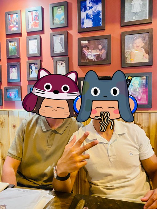 Two men sitting at a restaurant table in front of a wall of framed photos, with cartoon filter overlays covering their faces—one with a purple cat, the other with a gray cat eating noodles.