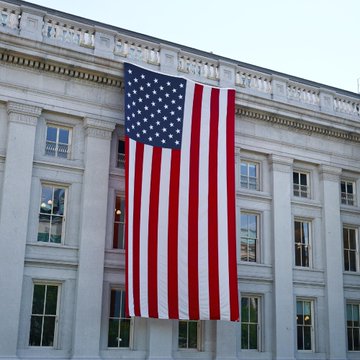 A massive American flag hangs from a stately government building, symbolically illustrating the US government's seizure and transfer of $297 million in Bitcoin and Ethereum to Coinbase Prime from three criminal forfeitures, as tracked by Arkham. The imposing federal architecture underscores the government's role in these high-stakes cryptocurrency asset movements.