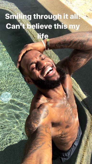 A shirtless man with tattoos smiles joyfully while relaxing in a pool, with text overlay reading 'Smiling through it all. Can't believe this my life'.
