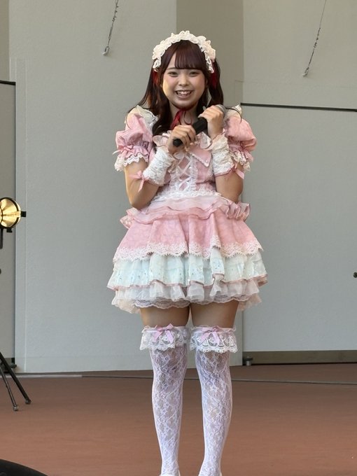 A performer in an elaborate pink and white frilly maid-style dress with a headband holds a microphone on stage, smiling at the camera.