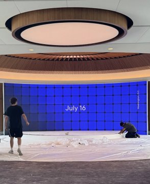 Two workers install a massive video wall displaying "July 16" in blue and white, fitting the "LOADING" theme as they prepare a large circular stage for an upcoming product launch event.