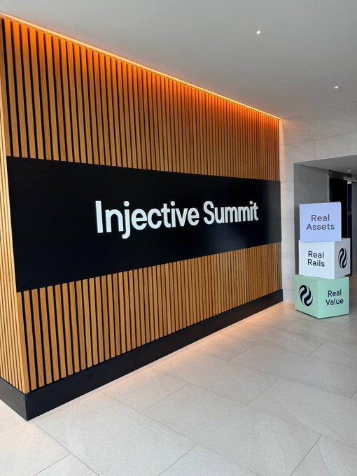 An 'Injective Summit' sign on a wood-paneled wall with illuminated white text, alongside stacked boxes reading 'Real Assets,' 'Real Rails,' and 'Real Value.'
