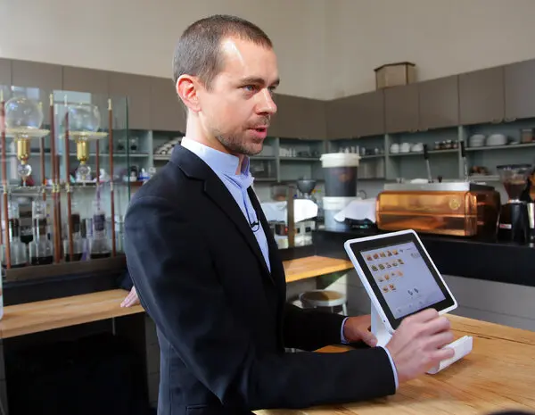 A man in a black suit and blue shirt standing at a wooden counter in what appears to be a café or restaurant, looking at a touchscreen POS ordering system.