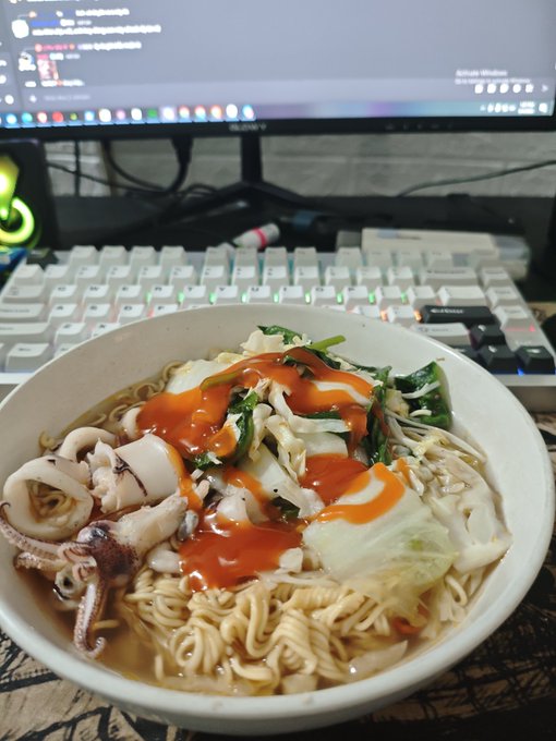 The image features a bowl filled with noodles topped with various ingredients such as vegetables and sauce. The bowl is placed next to a computer keyboard, suggesting that someone was working or studying while enjoying their meal. A person can be seen typing on the keyboard in the background, indicating they might have been using the computer for work or study purposes.