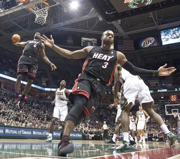 Miami Heat player number 3, Dwyane Wade, reaches for a loose basketball during an NBA game against the Milwaukee Bucks in their white jerseys.